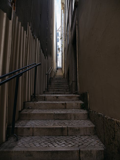 A narrow outdoor staircase with stone steps leading upward between two tall, enclosed walls—one on the left with a black metal railing and a temporary construction barrier, and one on the right with