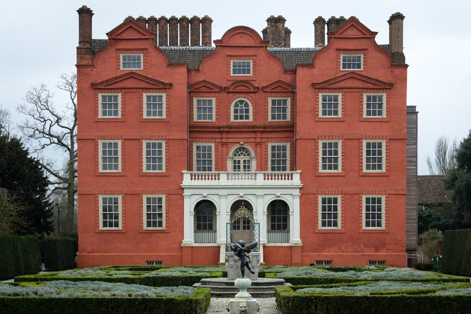 A large, historic red brick house with a symmetrical façade, featuring three stories and multiple evenly spaced rectangular windows with white frames. The central entrance has a white portico support