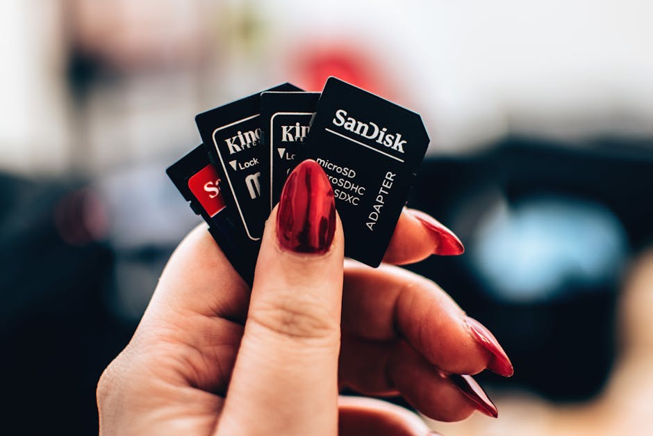 A close-up of a person's hand with long, red, manicured nails holding a small stack of digital storage cards, including various types of SD cards such as SanDisk, Kingston, and other brands, each with